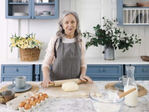 Mature woman with silver hair smiling in modern blue kitchen while baking with flour, eggs, and rolling pin on marble countertop