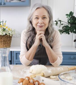 Mature woman with silver hair smiling in modern blue kitchen while baking with flour, eggs, and rolling pin on marble countertop